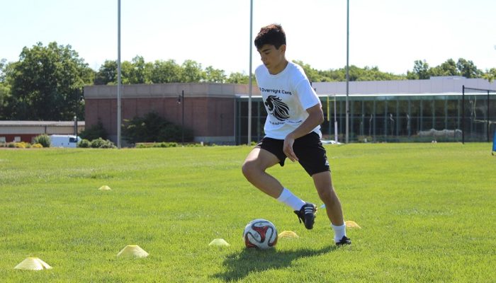 A teenage boy wearing a white shirt and black shorts completes soccer drills during practice