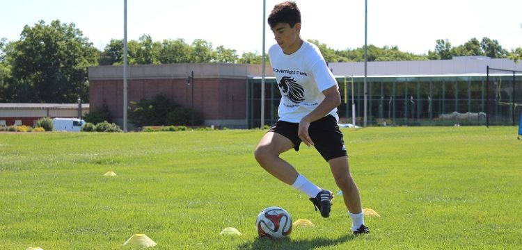 A teenage boy wearing a white shirt and black shorts completes soccer drills during practice