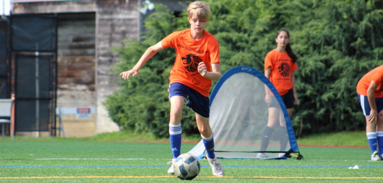 A teenage boy with blonde hair wearing an orange soccer kit dribbles a soccer ball away from a blue net