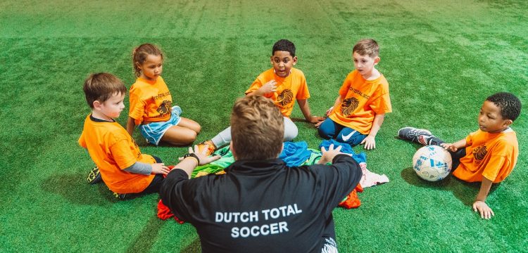 A youth soccer coach in a black jacket that says "Dutch Total Soccer" sitting in a circle with five youth soccer players wearing orange kits