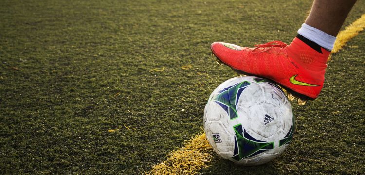 The foot of someone wearing a red Nike soccer cleat steps on a soccer ball