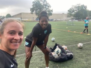 Three female soccer players posing for a selfie in front of a mesh bag filled with soccer balls