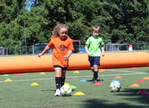 Two young soccer players, a girl in an orange shirt, and a boy in a lime green shirt, learn drills