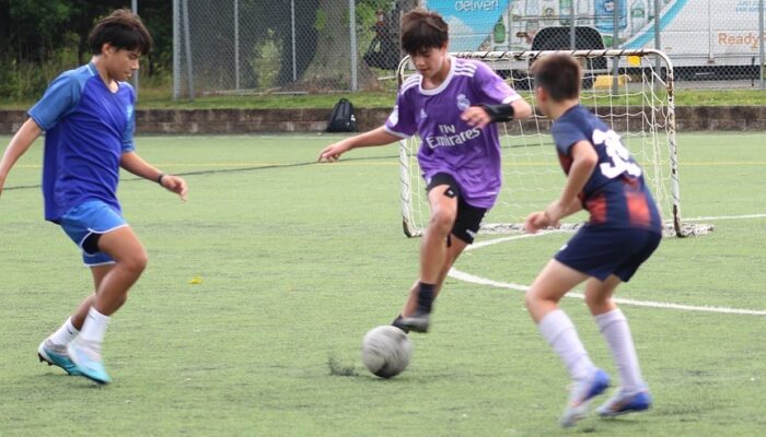 Three teenagers go after a soccer ball. One is wearing a blue kit, one is wearing a purple kit, and one is wearing a navy kit
