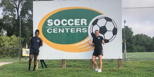 Two female soccer coaches stand in front of a white sign with a green and yellow circle that says "Soccer Centers" inside of it