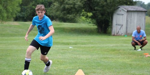 A teenage boy wearing a blue soccer kit dribbles upfield while a coach watches on