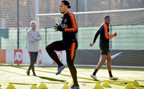 Two soccer players warm up while wearing black and orange track suits and a coach in a gray and black sweatsuit watches on